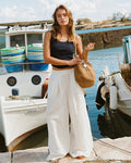 Lifestyle front view of a model wearing the LSPACE Ash Tank in midnight blue, a cropped ribbed racerback tank, styled with white wide-leg pants and a woven tote bag at a harbor setting.