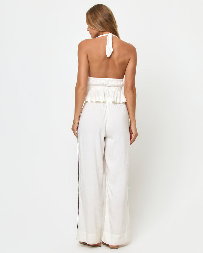 Back view of a model wearing the LSPACE August Top in cream on a white background, showing the open back, halter-neck tie, and peplum hem, styled with cream wide-leg pants.