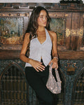Lifestyle front view of a model wearing the LSPACE Callie Halter in metallic silver, featuring a deep V-neckline with a twist-front detail, halter neck, and semi-sheer metallic knit fabric, styled with black wide-leg trousers and a crystal rhinestone clutch, photographed against an ornate wooden backdrop.
