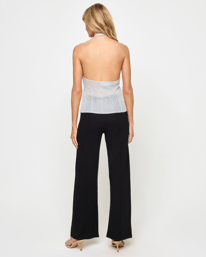Back view of a model wearing the LSPACE Callie Halter in metallic silver, showing the open back, halter neck tie, and metallic ribbed knit fabric, styled with black wide-leg flare trousers.