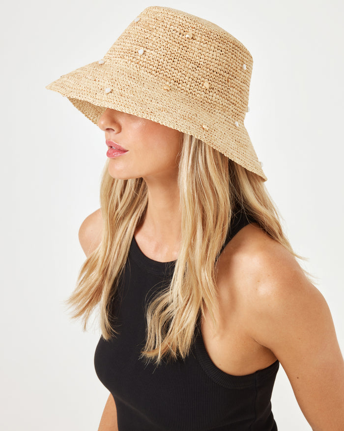 Side profile of model wearing the Carmel Island Hat in natural, a woven raffia bucket hat with pearl embellishments, paired with a black sleeveless top.