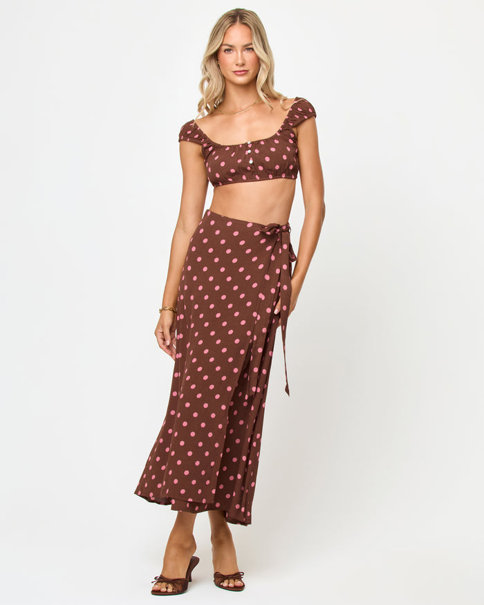 Full front view of a model wearing the LSPACE Ever Top in alotta polka dot, a brown and pink polka dot cap-sleeve crop top with button front detail, styled with the matching wrap midi skirt.