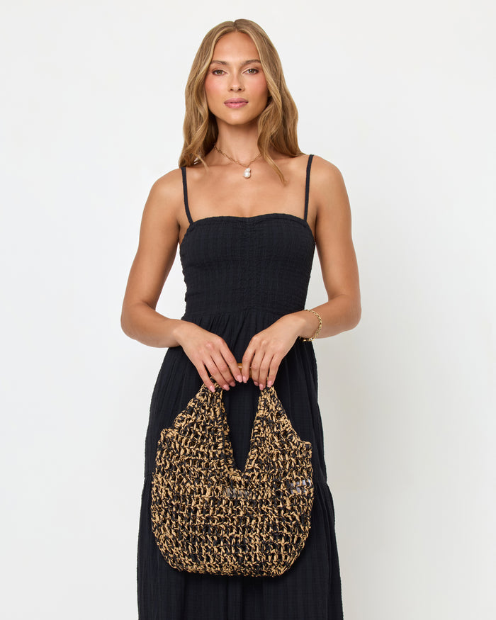 Front view of a model holding the natural and black Faye Bag with both hands, wearing a black maxi dress.