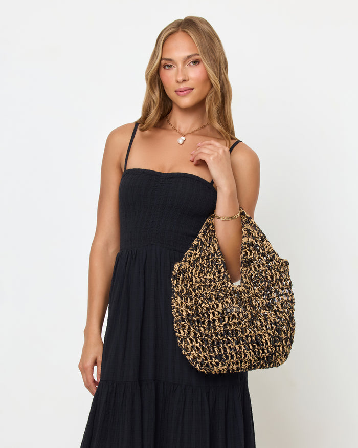 Side view of a model carrying the natural and black Faye Bag on her shoulder, wearing a black maxi dress.