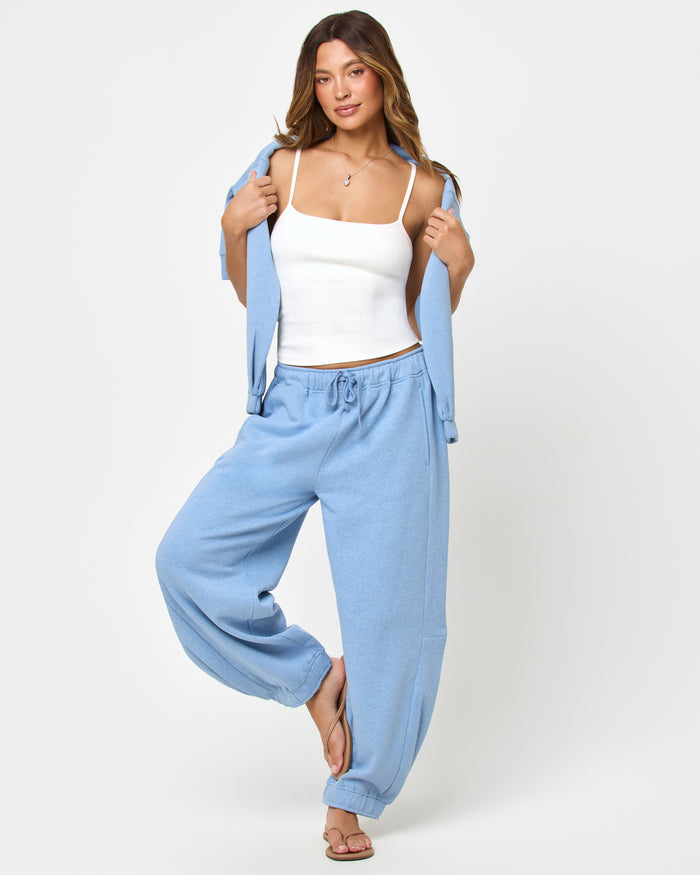 Front full-length studio view of the Fleur Pant in chambray blue, chambray blue wide-leg drawstring jogger pants with cuffed ankles, styled with a white spaghetti-strap tank and a chambray blue open-front cardigan held open, tan flip flops.