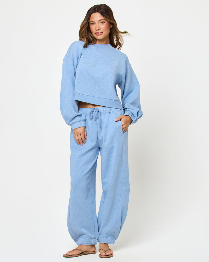 Front full-length studio view of the Fleur Sweatshirt and Fleur Pant in chambray blue, a chambray blue cropped crewneck sweatshirt paired with matching chambray blue wide-leg drawstring jogger pants and tan flip flops.
