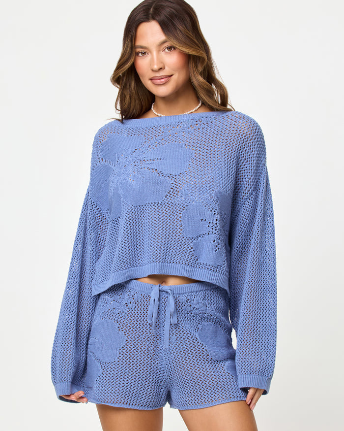Close-up of a model wearing the LSPACE Island Time Sweater in Chambray, styled with the matching Island Time Short, highlighting the open-knit crochet floral design and long sleeves.