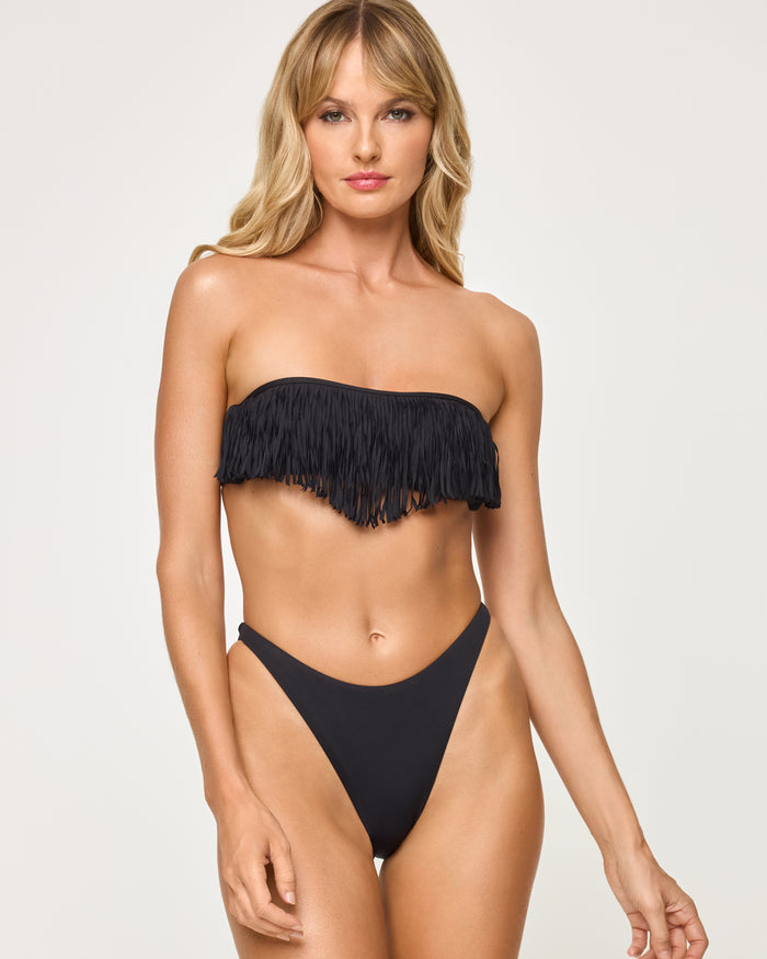Front view of a model wearing the LSPACE black Leilani Bikini Bottom, a smooth high-cut cheeky bikini bottom, paired with a matching black fringe bandeau bikini top on a neutral background.