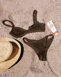 Flat lay of the espresso LSPACE Mackenzie Bikini Bottom and matching crochet mesh Tyla Bikini Top on sand, styled with a straw hat and sunscreen tube.