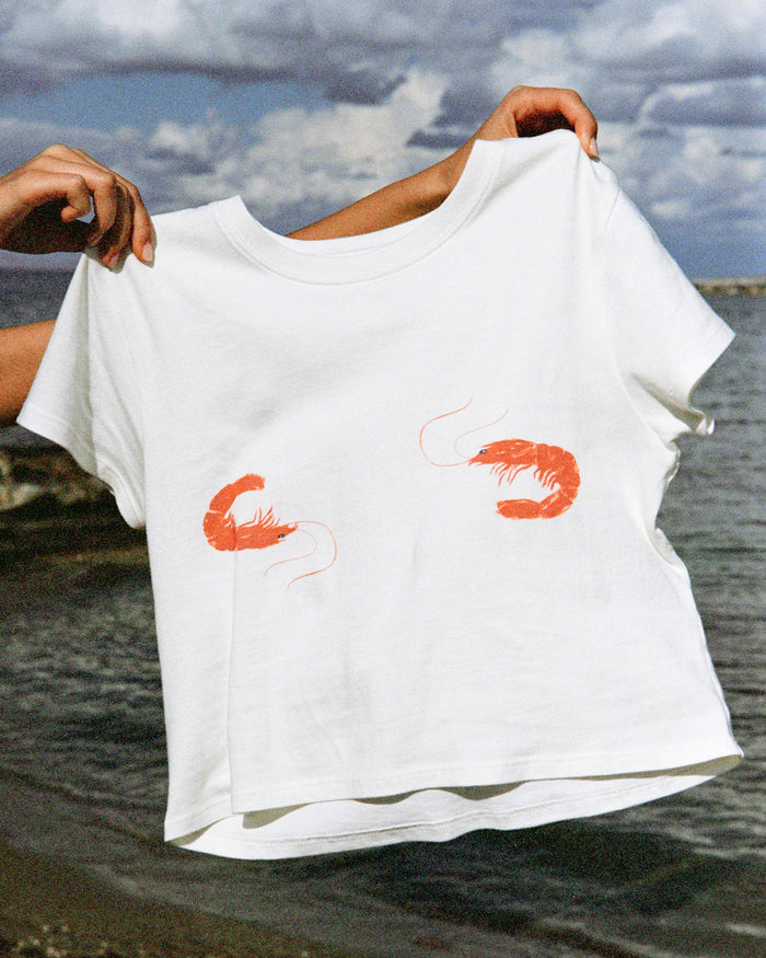 Off-model view of the LSPACE Merci Baby Tee in Shrimp, held up outdoors near the ocean to display the white cropped silhouette and two coral-red shrimp graphic prints on the front.