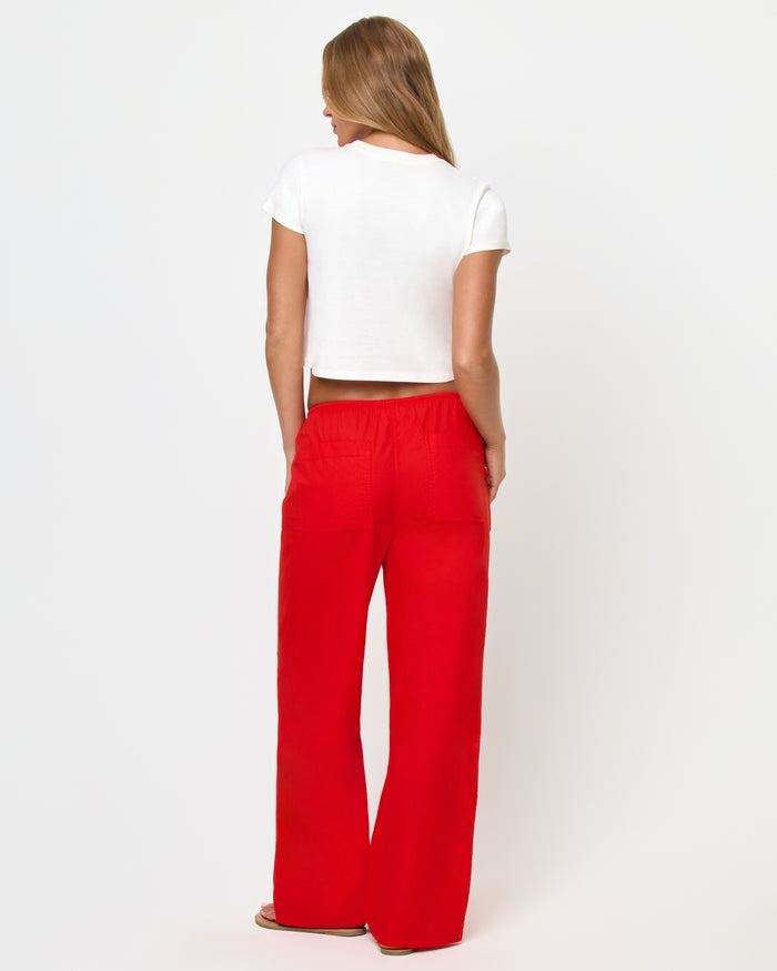 Back view of a model wearing the LSPACE Merci Baby Tee in Shrimp, showing the plain white cropped back, styled with red wide-leg drawstring pants.