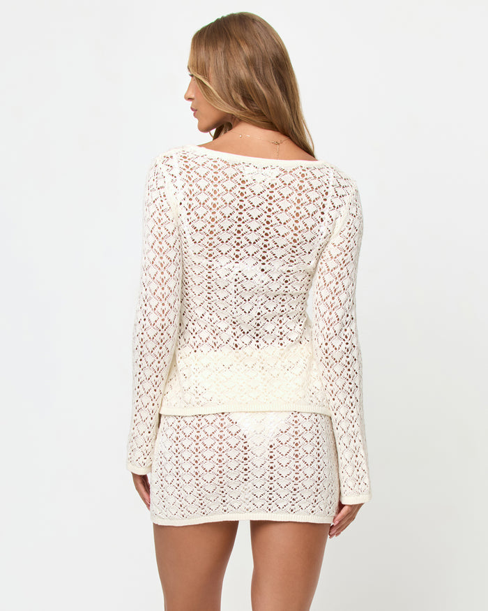 Back view of a model wearing the LSPACE Nova Top in cream, showing the long-sleeve crochet back with an open diamond lace pattern, styled with the matching LSPACE Nova Skirt.