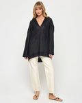 Front view of a model wearing the LSPACE Oasis Tunic in black, featuring an open V-neckline with collar, long sleeves, and a relaxed oversized silhouette, styled with cream wide-leg pants.