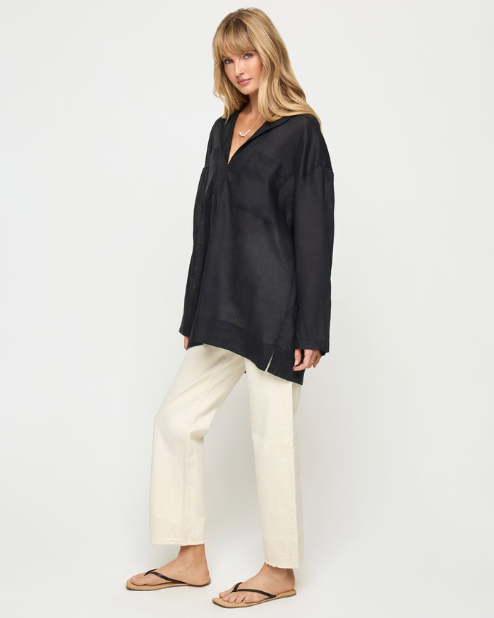 Three-quarter view of a model wearing the LSPACE Oasis Tunic in black, showing an open V-neckline, long sleeves, and a relaxed oversized silhouette, styled with cream wide-leg pants.