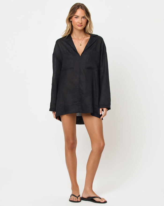Front view of a model wearing the LSPACE Oasis Tunic in black as a cover-up, featuring an open V-neckline with collar, long sleeves, and a relaxed oversized silhouette.