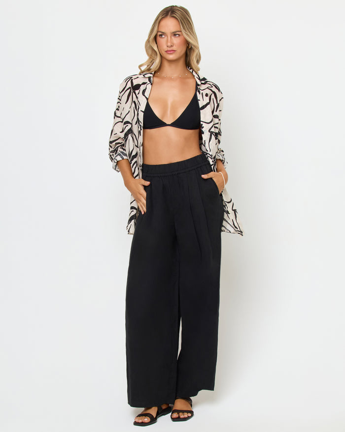 Front view of a model wearing the LSPACE Rio Tunic in heavy petal, an open-front linen button-up shirt with a cream and black bold botanical brushstroke print, styled over a black bikini top and wide-leg black pants.