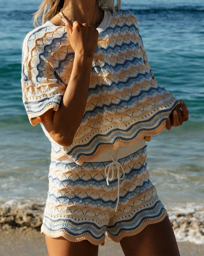 Close-up on model of the LSPACE Sunchaser Top in Daydreamer, a short-sleeve crochet top in a blue, peach, and white wave pattern with scalloped hem, styled as a set with the matching Sunchaser Short at the beach.