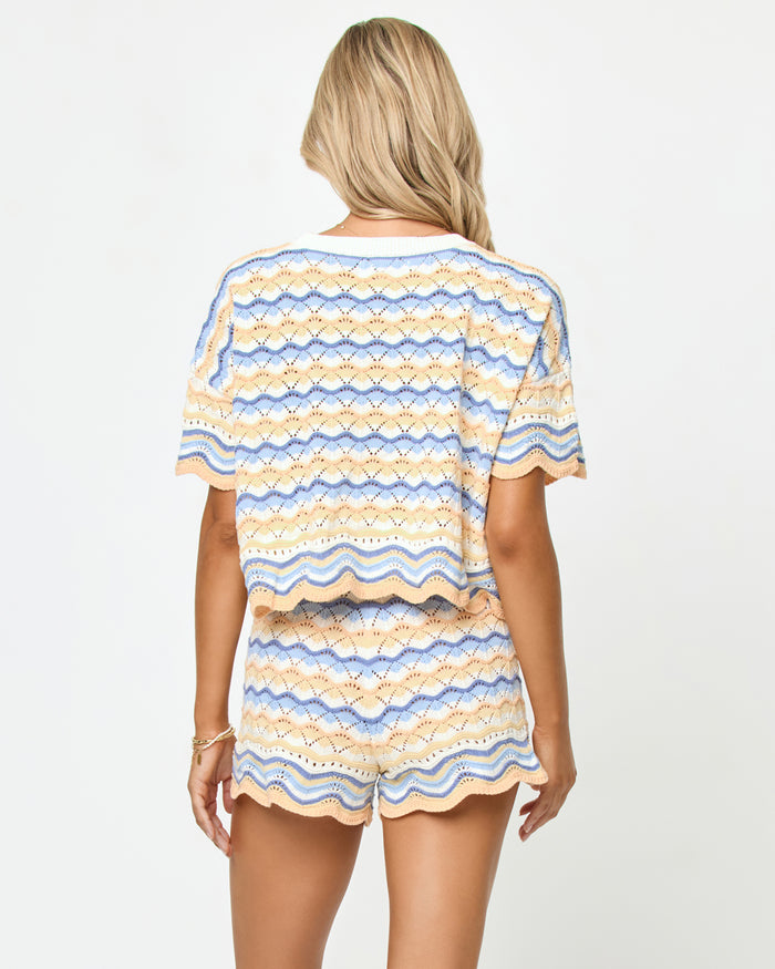 Back view on model of the LSPACE Sunchaser Top in Daydreamer, a short-sleeve crochet top in a blue, peach, and white wave pattern with scalloped hem, styled as a set with the matching Sunchaser Short.