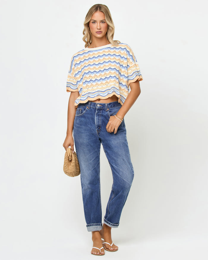 Front view on model of the LSPACE Sunchaser Top in Daydreamer, a short-sleeve crochet top in a blue, peach, and white wave pattern with scalloped hem, styled with blue denim jeans and a straw clutch.