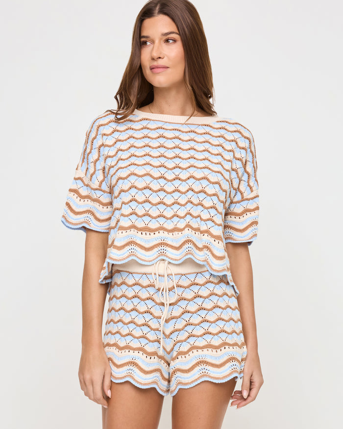 Angled front view of a model wearing the LSPACE Sunchaser Top in north shores, a boxy crochet knit top with a wave scallop pattern in light blue and tan, paired with the coordinating shorts.