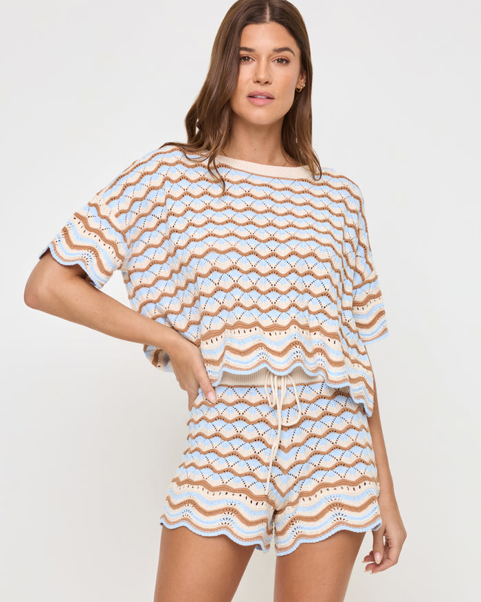 Front view of a model wearing the LSPACE Sunchaser Top in north shores, featuring a relaxed boxy fit, short sleeves, and a wave scallop crochet pattern in light blue and tan, styled with the matching shorts.