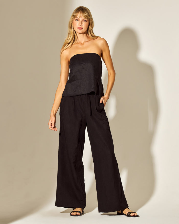 Front view of a model wearing the LSPACE Tessa Top in black, a strapless tube top with a straight neckline and peplum hem, styled with wide-leg black pants in a campaign-style editorial setting.