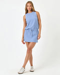 Front full-length studio view of the Upbeat Dress in chambray, a chambray blue ribbed sleeveless mini dress with a drawstring waist, styled with white Adidas sneakers.